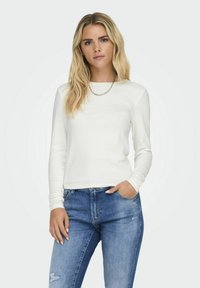 Chemise blanche à manches longues en tissu doux avec un col rond, associée à un jean bleu ajusté. Texture subtile, design épuré et sans motifs visibles.