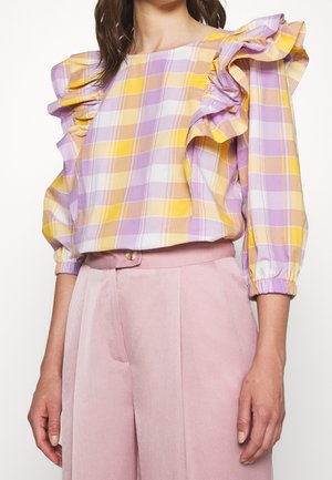 Vrouw draagt een geel, roze en lavendel geruit topje met ruches op de schouders en lichtroze hoge taille broek.