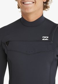 Traje de neopreno negro con cuello alto, costuras planas y un bolsillo con cremallera en el pecho con un pequeño tirador. El material tiene una textura suave.