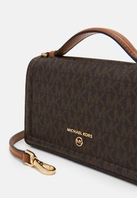 Ruskea kangaskäsilaukku, jossa on kultaiset yksityiskohdat, logolla ja irrotettavalla ruskealla hihnalla. Pintakuvio, jossa on keskenään lomittain olevia MK-kirjaimia.