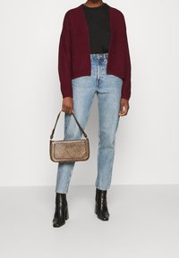 Burgundy stickad kavaj över en svart topp, matchad med ljusblå jeans och svarta klackboots. Håller en mönstrad brun axelväska.