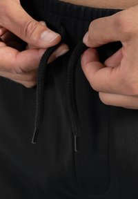 Zwarte joggings met een gevlochtenkoord en plastic eindstukjes. Handen grijpen de koorden tegen een textuur achtergrond.