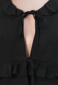 Black garment with a ruffled collar, featuring a V-neckline and ties at the front. The fabric appears soft with a smooth texture.