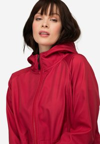 Veste légère rouge à capuche avec fermeture éclair sur le devant, présentant une texture lisse et des détails de couture. La capuche est réglable avec un cordon de serrage.