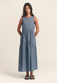 Mėlyna ilga be rankovių suknelė su sluoksniuotu dizainu, apvalia apykakle ir šoninėmis kišenėmis. Pagaminta iš lengvos, lygaus audinio tekstūros medžiagos.