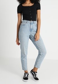 Svart kortärmad topp med knappknäppning, ljusblå högmidjade jeans och svarta sneakers med vita sulor. Enfärgad bakgrund.