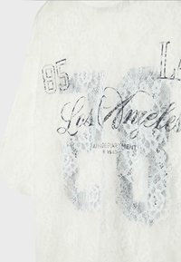 Tissu en dentelle blanche avec texte en écriture noire "Los Angeles" et chiffres "85" superposés sur un motif floral en dentelle.