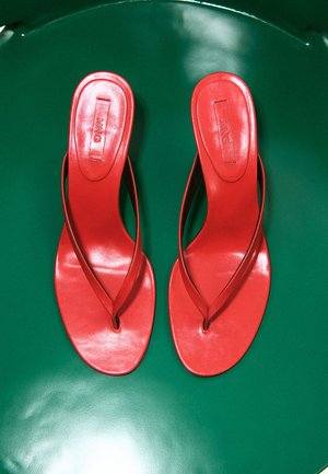 Para czerwonych sandałów na stopę typu slip-on z cienkimi paskami, ułożonych obok siebie na gładkiej, ciemnozielonej powierzchni.