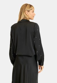 Czarna bluzka z długim rękawem, zdobiona perłowymi akcentami na rękawach. Gładki materiał o luźnym kroju i klasycznym fasonie kołnierzyka.