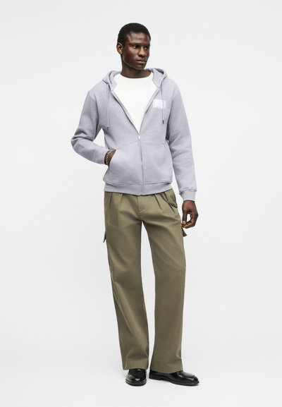 Άνδρας που στέκεται φορώντας ανοιχτόγκρι φούτερ με φερμουάρ, λευκό μπλουζάκι, χακί παντελόνι cargo, μαύρα παπούτσια και βραχιόλι στον αριστερό καρπό.