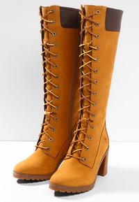 Botas altas de ante en color marrón claro con un cierre de cordones en la parte delantera, ojales de metal, acentos en cuero marrón y un tacón robusto. Superficie texturizada y suela con relieve.