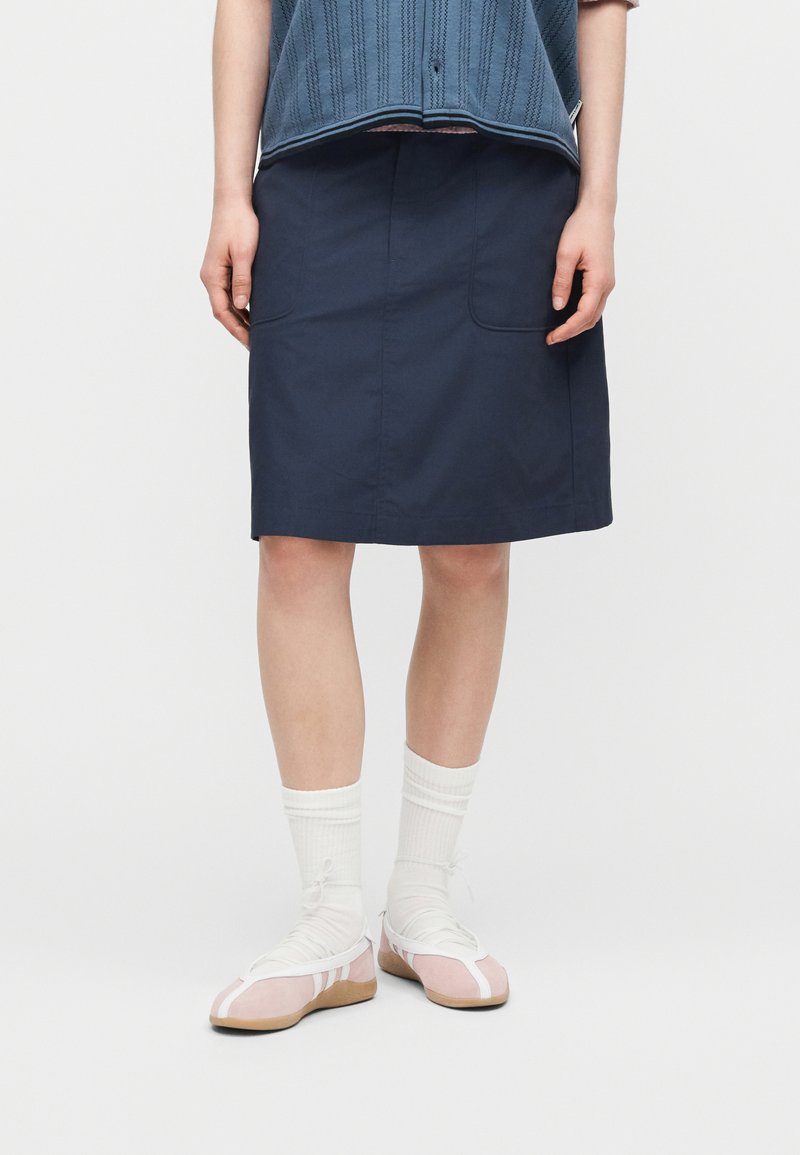 Persona con una gonna blu navy al ginocchio, calzini bianchi e ballerine rosa con cinturini bianchi, in piedi davanti a uno sfondo semplice.