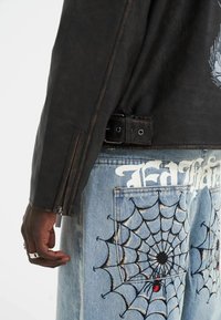 Black leather jacket with a side zipper. Blue denim pants featuring embroidered spider webs and bold white text on the back pockets.