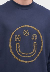 Sudadera de color navy con un diseño de cara sonriente de alambre de espino amarillo que presenta las letras H, G y O dentro del contorno circular.