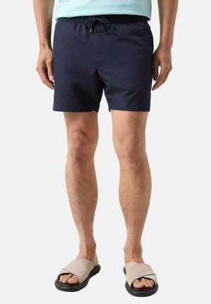 Mężczyzna ubrany w granatowe szorty ze ściągaczem i beżowe klapki z czarną podeszwą stoi na białym tle.