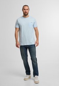 T-shirt azzurro chiaro a maniche corte con taschino sul lato sinistro, abbinato a jeans scuri e sneakers bianche. Il tessuto appare morbido e informale.