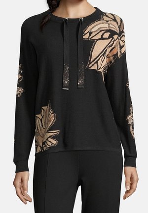 Haut noir à manches longues avec de grands motifs de feuilles beiges sur l'épaule, la manche et l'ourlet, doté d'un col rond avec des cordons noirs.