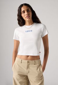 T-shirt bianca cropped in cotone con collo rotondo e logo blu Levi's sul petto, abbinata a pantaloni beige.