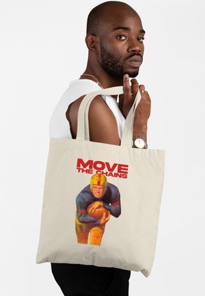 Muž držící béžovou tašku tote s ilustrací vintage fotbalového hráče a červeným nápisem "POSUNOUT ŘETĚZY" na bílém pozadí.