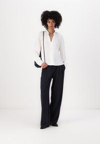 Blusa bianca a maniche lunghe con scollatura a V, abbinata a pantaloni larghi blu navy. La modella tiene una borsa a tracolla nera. Tessuto morbido e leggero.