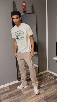 Vit t-shirt med grön text, beige byxor och grå sneakers. Står framför en spegel, med en neutral vägg och trägolv.