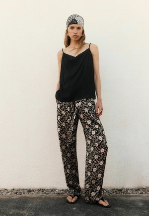 Femeie purtând o camisolă neagră, pantaloni largi cu model floral, șlapi negri și un batic alb cu model, stând în fața unui perete simplu.