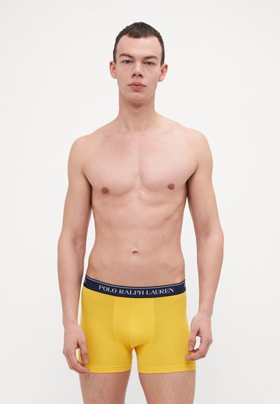 Rumene bokserice s temnim pasom, na katerem je napis "POLO RALPH LAUREN". Gladek material, prilegajoča oblika in dolžina do sredine stegen.