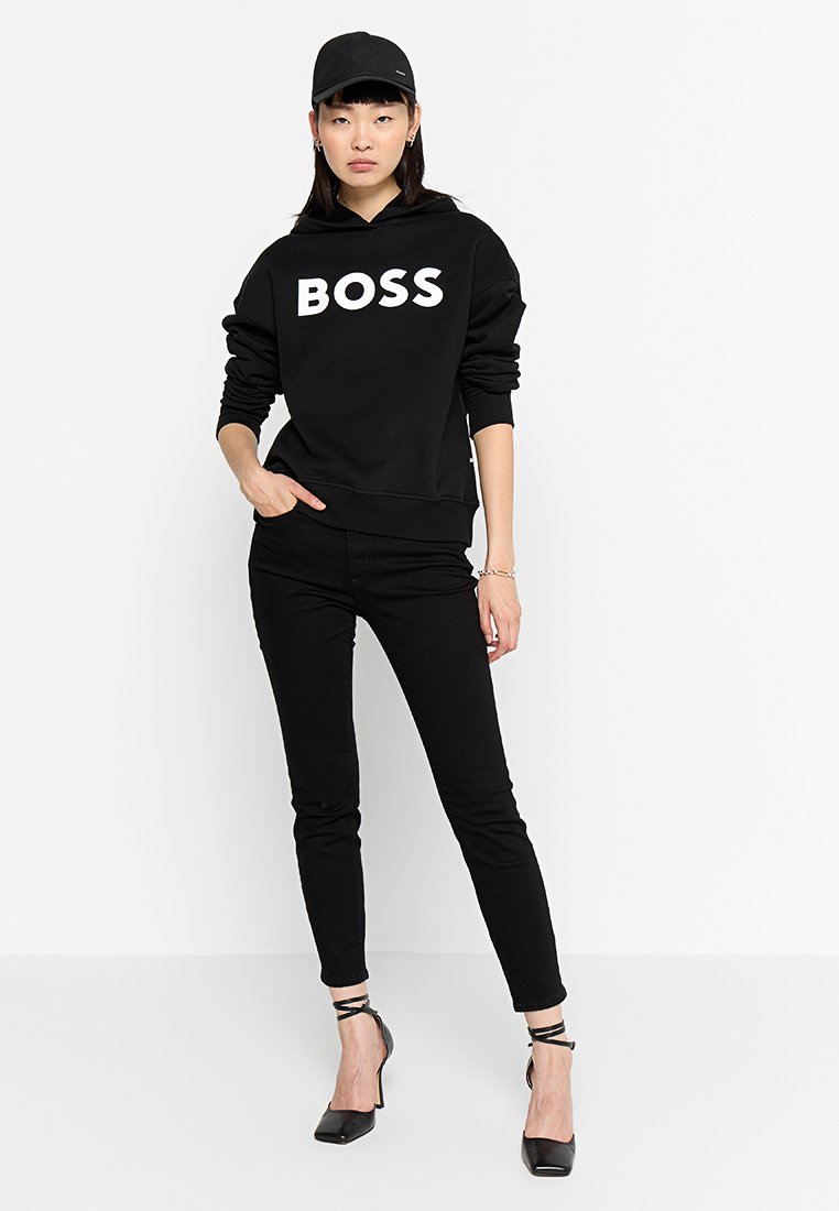 Zwarte hoodie met "BOSS" in vetgedrukte witte tekst, gecombineerd met zwarte skinny jeans en zwarte strappy heels, gedragen met een zwarte pet.