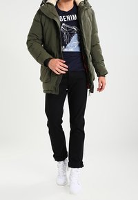 Oliwkowa pikowana kurtka z kapturem wyściełanym futrem, zestawiona z granatowym T-shirtem z nadrukiem, czarnymi spodniami i białymi trampkami.