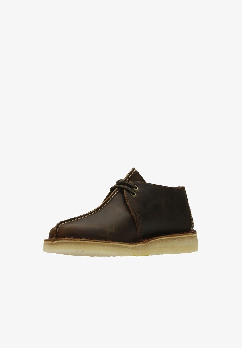 Brown leather desert boots with a round toe, contrasting white stitching, and a light-colored rubber sole. Unique design with minimal hardware.