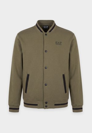 Un homme porte une veste universitaire vert olive, une chemise et un pantalon noirs, une casquette blanche et des chaussures noires, et tient un grand sac cabas noir.