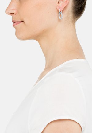 Boucles d'oreilles en argent en forme d'ovale, avec une finition texturée, suspendues à un petit crochet. Portées par un mannequin à la peau claire.