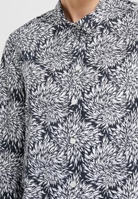 Camicia button-up in cotone blu navy con motivo floreale bianco; presenta un colletto a punta e bottoni frontali; texture leggera e liscia.