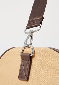 Sac en toile beige avec une sangle marron, présentant un clip en métal, des accents en cuir et une surface texturée. Gros plan sur le détail de l'attache.