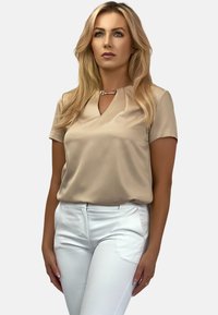 Beżowa bluzka z krótkim rękawem i dekoltem w kształcie litery V, z metalowym akcentem przy dekolcie, zestawiona z dopasowanymi białymi spodniami. Gładka faktura materiału.