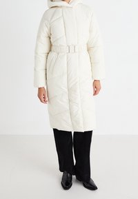 Puffy cream-colored long coat with a belted waist, hood, and diagonal quilted pattern. Worn over black corduroy pants and shoes.
