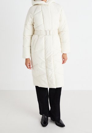 Puffy cream-colored long coat with a belted waist, hood, and diagonal quilted pattern. Worn over black corduroy pants and shoes.