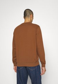 Sweat-shirt marron avec col rond et manches longues, présentant des poignets et un ourlet côtelés. Le tissu a une texture lisse et une légère élasticité.