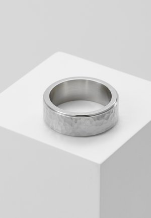 Bague large en argent avec texture martelée reposant sur un socle cube blanc sur un fond gris clair.