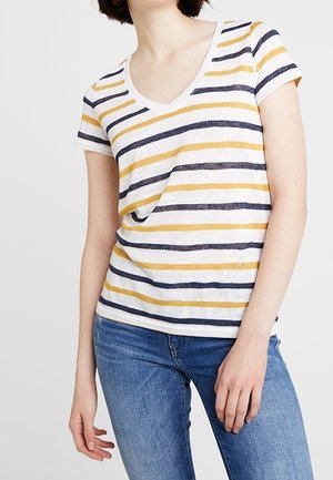 Vrouw draagt een wit T-shirt met V-hals en horizontale marineblauwe en mosterdkleurige strepen, gecombineerd met een blauwe jeans, staand tegen een egale achtergrond.
