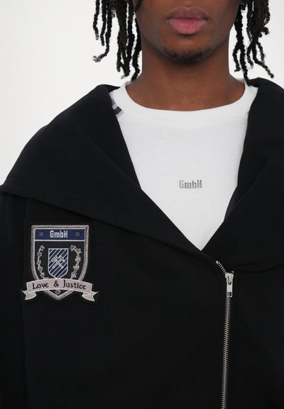 Jachetă neagră cu fermoar și o glugă mare, având un patch brodat care scrie "GmbH" și "Dragoste & Justiție". Sub ea, un tricou alb cu guler rotund.