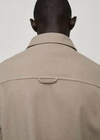 Impermeabile beige con colletto classico, caratterizzato da una texture liscia e un discreto dettaglio con pattina sul retro, adatto per abbigliamento casual o formale.