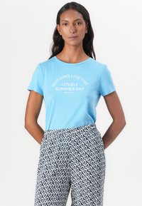T-shirt blu a maniche corte con testo bianco "NIENTE È SIMILE A QUESTO SPLENDIDO GIORNO ESTIVO" abbinato a pantaloni a fantasia chiara e scura.