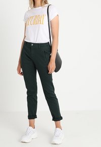 T-shirt bianca con la scritta 'SABATO' in giallo, pantaloni verdi scuri, sneakers bianche e una borsa a tracolla nera; stile casual con i pantaloni arrotolati.