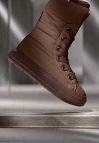 Brun puffer-lignende sneaker med polstret overdels, rund tå, snørebånd foran og strukturert gummisåle. Detaljene inkluderer kontrasterende snorer og sømmer.