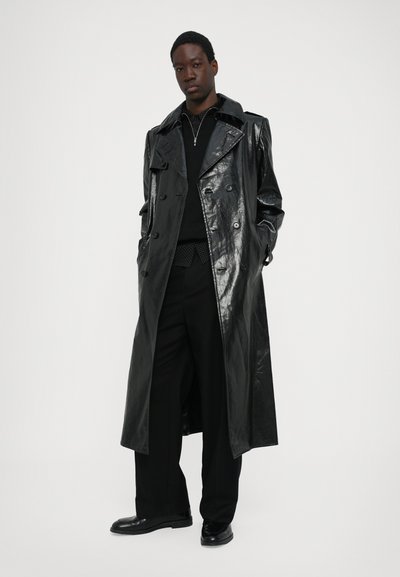 Abrigo de cuero negro con acabado brillante, botones de doble botonadura, solapas anchas y bolsillos laterales. Lleva puesto un suéter negro y pantalones.