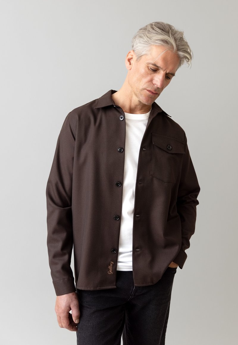 Camicia button-up marrone con colletto, caratterizzata da una tasca sul petto e un dettaglio ricamato "Gallery". Indossata sopra una maglietta bianca.