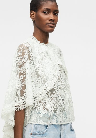 Femme portant un haut blanc en dentelle à motif floral avec un col montant à volants et des manches mi-longues, associé à un jean bleu clair sur un fond uni.