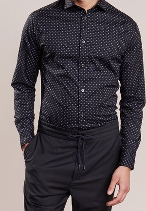 Homme portant une chemise noire à boutons avec un petit motif triangulaire bleu et un pantalon noir à cordon, mains dans les poches, debout devant un fond uni.