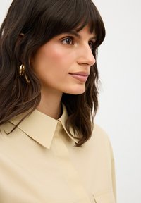 Camicia button-up beige chiaro con colletto, caratterizzata da una texture liscia e due tasche frontali. I lunghi capelli scuri incorniciano delicatamente il viso.